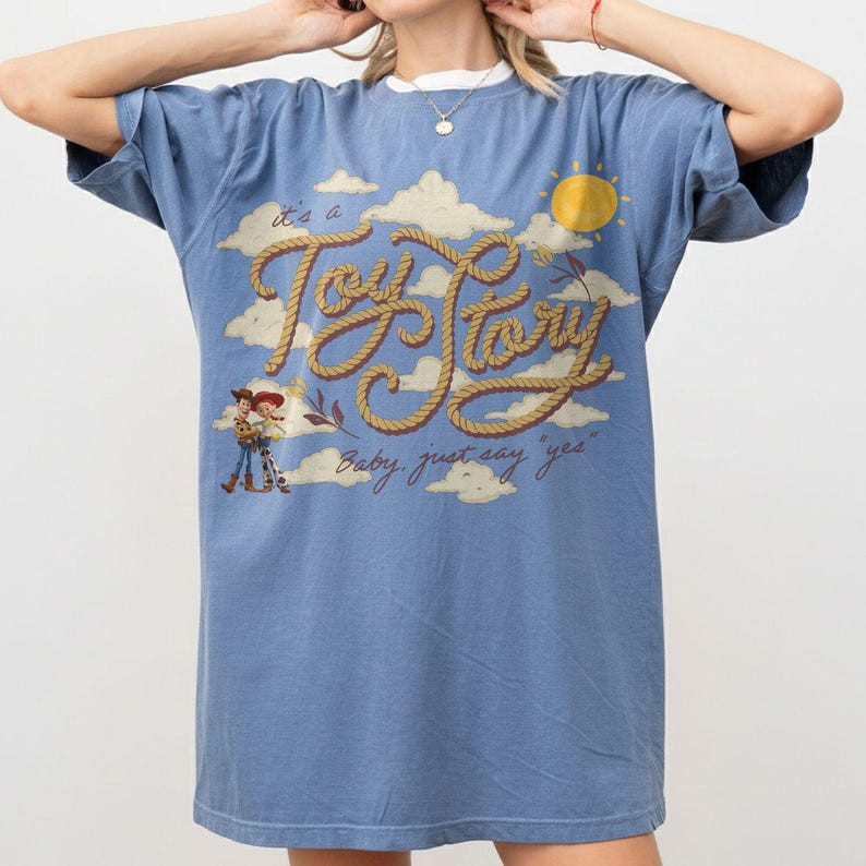 Op de afbeelding: Een blauw t-shirt met korte mouwen voorzien van een afbeelding van wolken, een gele zon en de tekst "it's a Toy Story" in een touwachtig lettertype. Onder de tekst toont het t-shirt de zin "Baby, just say "yes"" en twee stripfiguren.
