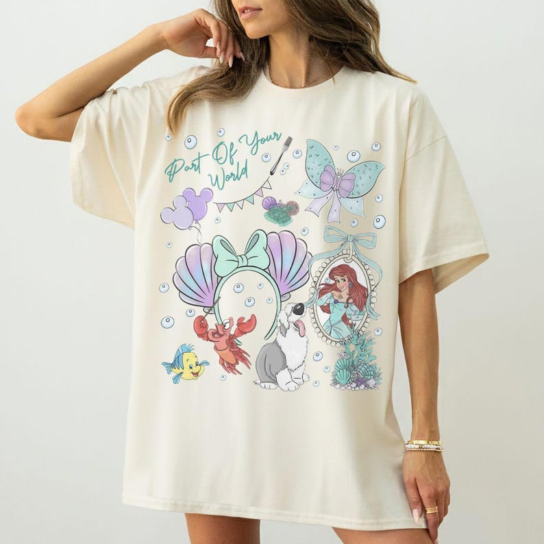 May include: An off-white t-shirt featuring a graphic print with the text Part Of Your World. The design includes a mermaid, a crab, a small yellow fish, a dog, a bow, a seashell headband, and bubbles in shades of blue, purple, and red.