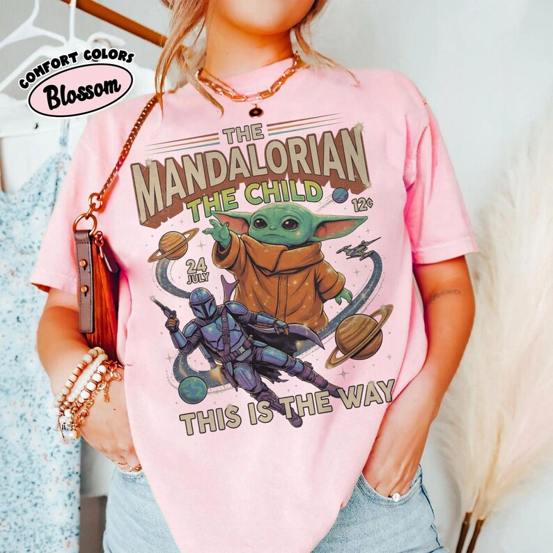 Op de afbeelding: Een lichtroze t-shirt met een grafische print van The Mandalorian en The Child. Het ontwerp bevat ruimte-elementen, planeten en de tekst "The Mandalorian", "The Child", "24 July", "12c" en "This is the way" in een vintage stijl.