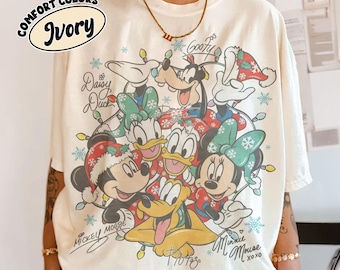 May include: An ivory t-shirt featuring a graphic print of Disney characters including Mickey Mouse, Minnie Mouse, Donald Duck, Daisy Duck, Goofy, and Pluto wearing holiday attire. The design includes Christmas lights, snowflakes, and character names.