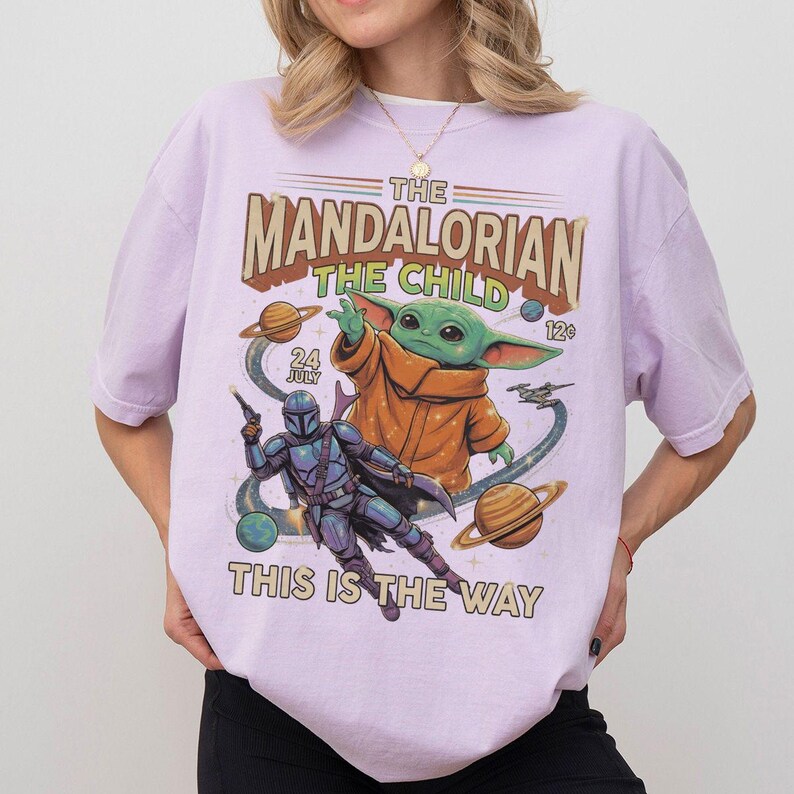 Op de afbeelding: Een lichtpaars t-shirt met een grafische print met de tekst THE MANDALORIAN THE CHILD 24 JULY 12c THIS IS THE WAY. Het ontwerp bevat een groen personage in een oranje gewaad, een blauw gepantserde figuur, planeten en een klein ruimteschip.