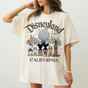 May include: Cream-colored t-shirt featuring a Disneyland graphic. The design includes the Disneyland logo, the text "Est.1955", and cartoon characters like Mickey and Minnie Mouse. The word "CALIFORNIA" is printed at the bottom of the graphic.