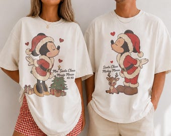 May include: Two off-white t-shirts featuring vintage-style Christmas graphics. One shirt displays Minnie Mouse as Mrs. Santa Claus with a tree and the text "Mrs Santa Claus Minnie Mouse XO XO". The other shows Mickey Mouse as Santa Claus with a reindeer.
