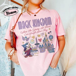 May include: A light pink short-sleeve t-shirt featuring a graphic print with the text MAGIC KINGDOM Where The Young At Heart Can Laugh And Play. The design includes a wizard mouse, a castle, a train, and various park-themed icons in purple.