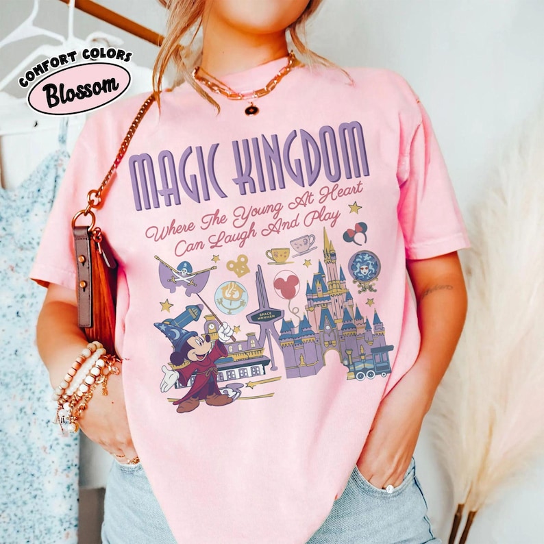 May include: A light pink short-sleeve t-shirt featuring a graphic print with the text MAGIC KINGDOM Where The Young At Heart Can Laugh And Play. The design includes a wizard mouse, a castle, a train, and various park-themed icons in purple.