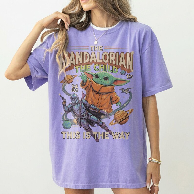 Op de afbeelding: Een lichtpaars t-shirt met een grafische print van The Mandalorian en The Child. Het ontwerp bevat planeten, sterren en de tekst "The Mandalorian The Child 24 July 12c This Is The Way" in een vintage stripboekstijl.