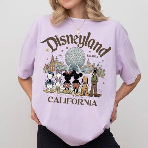 May include: A light purple t-shirt featuring the Disneyland logo with a graphic of Disney characters and the text "CALIFORNIA". The design includes Mickey and Minnie Mouse, Donald Duck, and Goofy. The shirt has short sleeves.