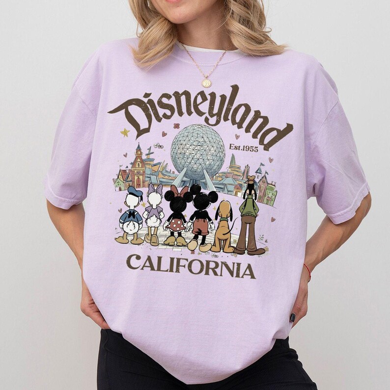 May include: A light purple t-shirt featuring the Disneyland logo with a graphic of Disney characters and the text "CALIFORNIA". The design includes Mickey and Minnie Mouse, Donald Duck, and Goofy. The shirt has short sleeves.