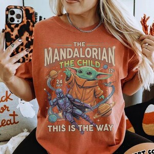 Op de afbeelding: Een roestkleurig t-shirt met een grafische print van The Mandalorian en The Child in de ruimte. Het ontwerp bevat planeten, sterren en de tekst "The Mandalorian The Child 24 July 12c This Is The Way" in een vintage lettertype.