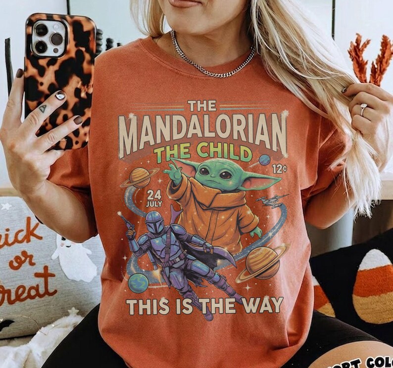 Op de afbeelding: Een roestkleurig t-shirt met een grafische print van The Mandalorian en The Child in de ruimte. Het ontwerp bevat planeten, sterren en de tekst "The Mandalorian The Child 24 July 12c This Is The Way" in een vintage lettertype.