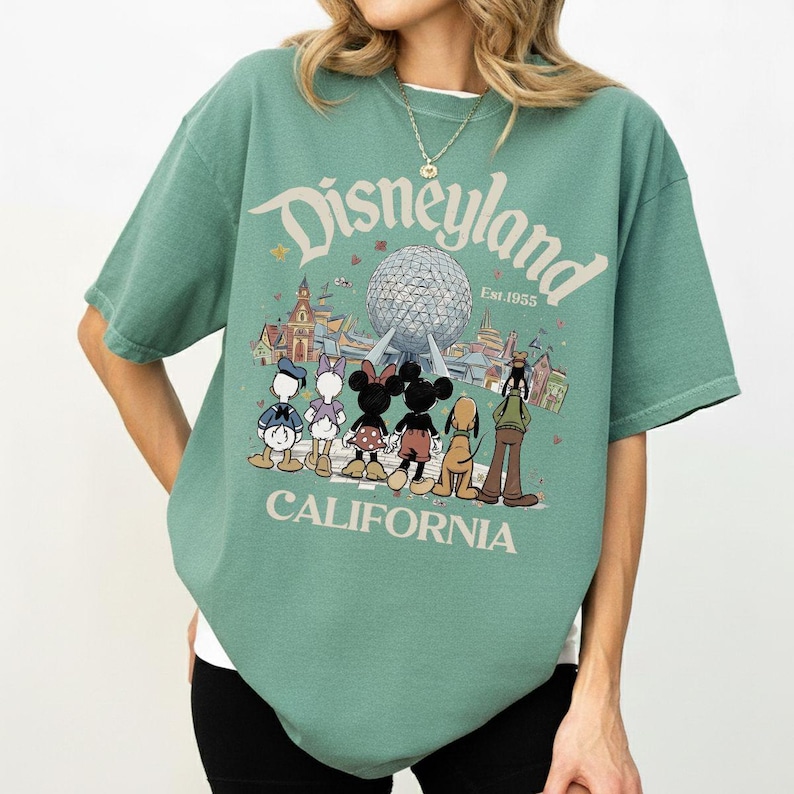 May include: Green t-shirt featuring the word "Disneyland" above a graphic of the Epcot ball and Disney characters. The text "Est. 1955" and "California" are also printed on the shirt. The shirt has short sleeves and a crew neck.