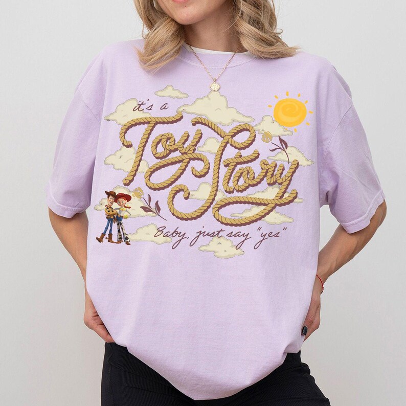 Op de afbeelding: Een lichtpaars t-shirt met korte mouwen en een grafische print van de tekst "it's a Toy Story" geschreven in een touwstijl lettertype, vergezeld van illustraties van Woody en Jessie, gele wolken, een zon en de tekst "Baby, just say "yes""