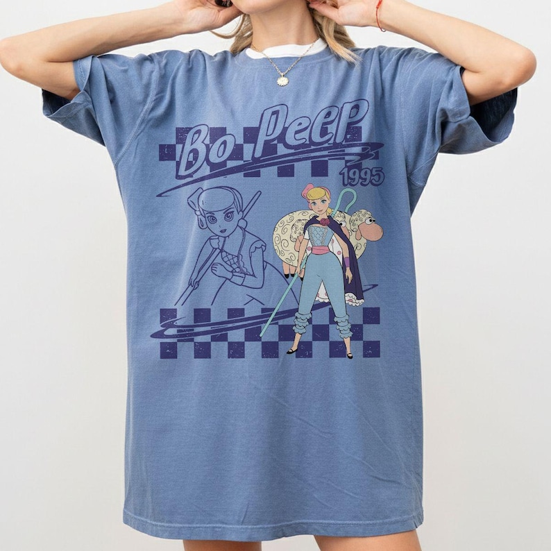 May include: A blue short-sleeve t-shirt featuring a graphic print of Bo Peep from 1995. The design includes a dark blue checkered pattern, a line art sketch of the character, and a full-color illustration of Bo Peep with a sheep and a shepherd's crook.