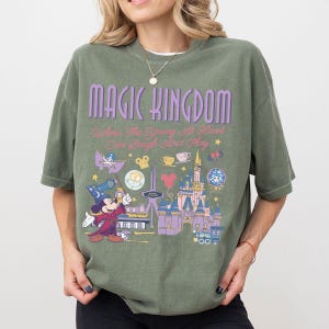 May include: An olive green short-sleeve t-shirt featuring the text MAGIC KINGDOM and Where The Young At Heart Can Laugh And Play. The graphic includes a wizard mouse, a castle, and various small icons in shades of purple, yellow, and red.