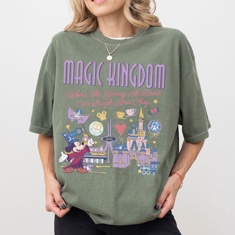 May include: An olive green short-sleeve t-shirt featuring the text MAGIC KINGDOM and Where The Young At Heart Can Laugh And Play. The graphic includes a wizard mouse, a castle, and various small icons in shades of purple, yellow, and red.