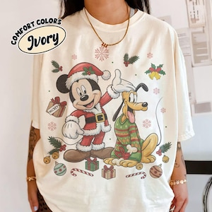 May include: An ivory t-shirt featuring a graphic of Mickey Mouse in a Santa suit and Pluto wearing a green patterned sweater. The design includes Christmas elements like snowflakes, holly, gift boxes, and ornaments. Text reads COMFORT COLORS IVORY.