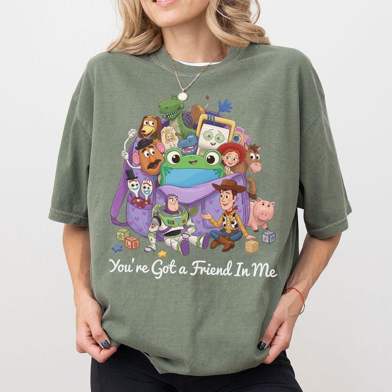 May include: An olive green t-shirt featuring a graphic print of various Toy Story characters emerging from a purple backpack. The design includes Buzz Lightyear, Woody, and other figures, with the text "You're Got a Friend In Me" printed below.