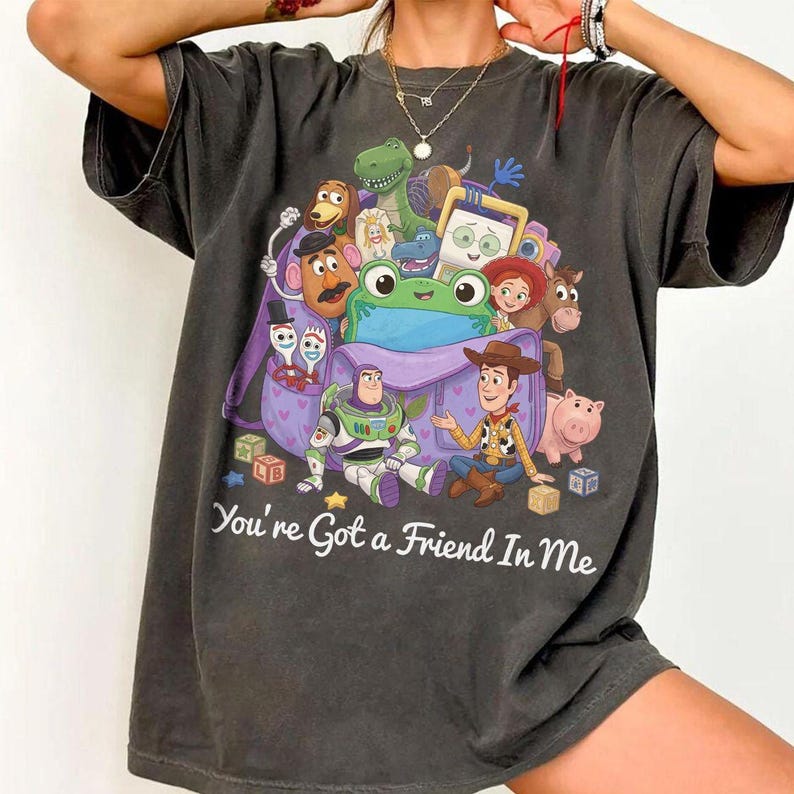 May include: A charcoal gray t-shirt featuring a graphic print of various animated toy characters gathered around a purple backpack. The text "You're Got a Friend In Me" is printed in white script below the characters. The shirt has short sleeves.