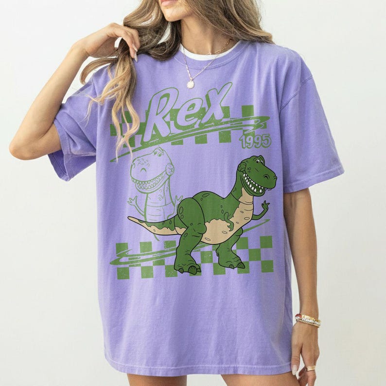May include: A light purple short-sleeve t-shirt featuring a green graphic of a Tyrannosaurus Rex. The design includes the text Rex and 1995, set against a green checkered background pattern. The shirt has a crew neckline and a relaxed fit.