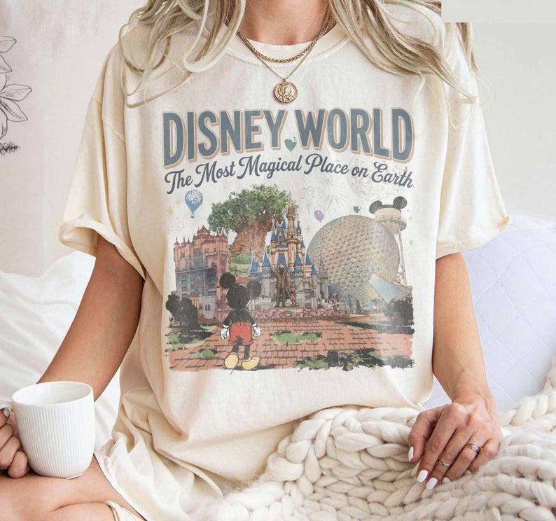 Puede incluir: Camiseta color crema con el texto "DISNEY WORLD The Most Magical Place on Earth" en azul y blanco. El gr&aacute;fico incluye ilustraciones de lugares emblem&aacute;ticos de Disney y Mickey Mouse. La camiseta tiene mangas cortas.