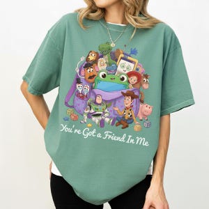 May include: A sage green short-sleeve t-shirt featuring a graphic print of various animated toy characters emerging from a purple backpack. The text "You're Got a Friend In Me" is printed in white below the graphic. The shirt has a crew neckline.