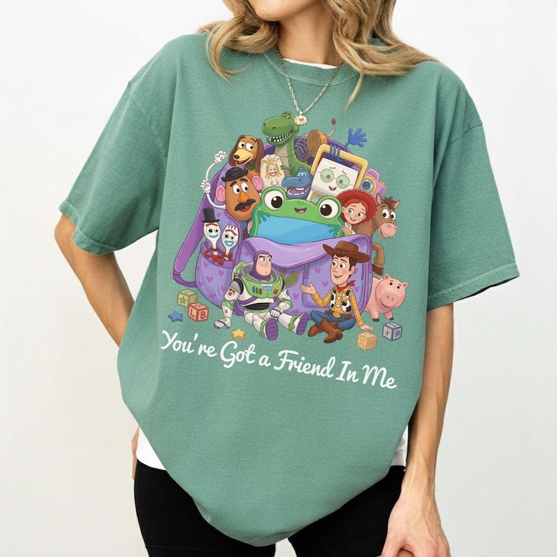 May include: A sage green short-sleeve t-shirt featuring a graphic print of various animated toy characters emerging from a purple backpack. The text "You're Got a Friend In Me" is printed in white below the graphic. The shirt has a crew neckline.