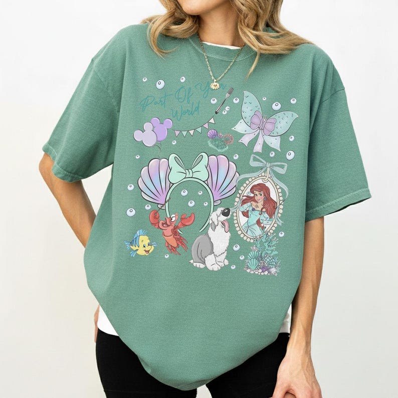 May include: A sage green short-sleeve t-shirt featuring a graphic print with the text Part Of Your World. The design includes a mermaid, a crab, a fish, a dog, seashell ears, a butterfly, and bubbles in shades of purple, blue, and white.