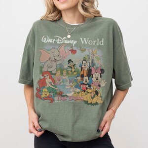 Puede incluir: Una camiseta verde oliva con un estampado gr&aacute;fico de varios personajes de Disney, incluidos Dumbo, Ariel, Cenicienta, Mickey Mouse, Minnie Mouse, el Pato Donald, Goofy y Pluto. El texto dice Walt Disney World y Magic Kingdom.