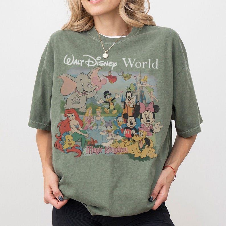 Puede incluir: Una camiseta verde oliva con un estampado gr&aacute;fico de varios personajes de Disney, incluidos Dumbo, Ariel, Cenicienta, Mickey Mouse, Minnie Mouse, el Pato Donald, Goofy y Pluto. El texto dice Walt Disney World y Magic Kingdom.
