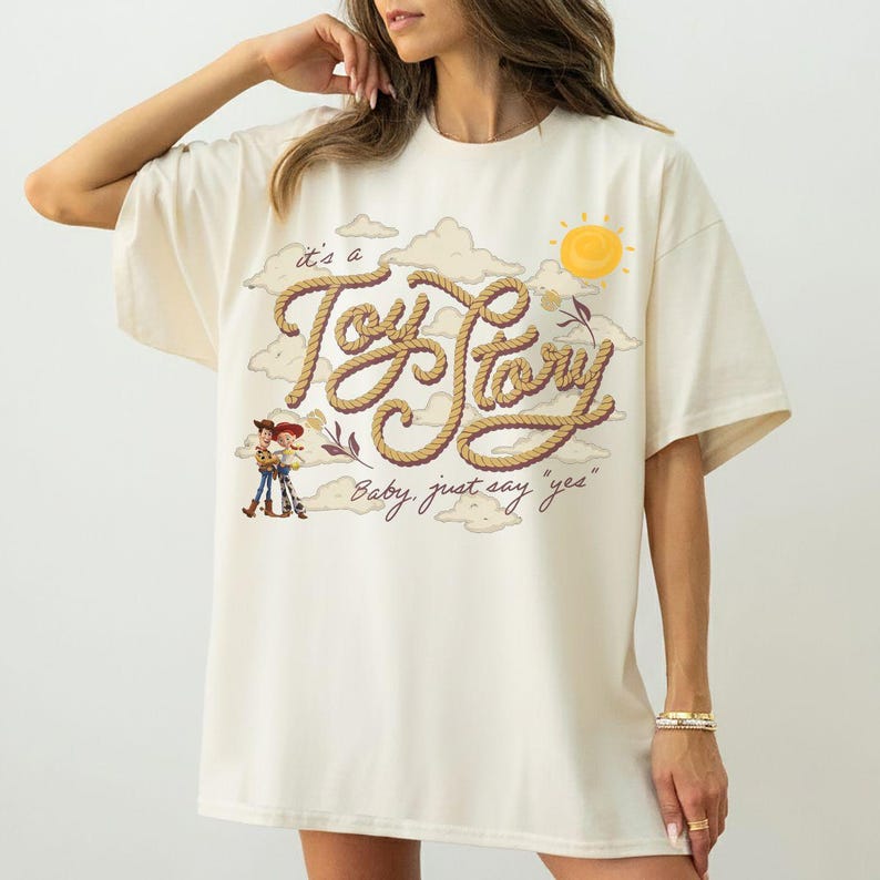 Op de afbeelding: Een off-white t-shirt met een grafische print van touwachtige letters die "it's a Toy Story" vormen. Onder de tekst staat de zin "Baby, just say 'yes'" gedrukt. Het ontwerp bevat stripfiguren, wolken en een gele zon.