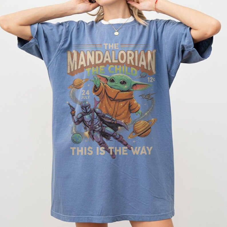 Op de afbeelding: Een blauw t-shirt met korte mouwen en een grafische print van The Mandalorian en The Child. Het ontwerp bevat planeten, sterren en de tekst THE MANDALORIAN THE CHILD 24 JULY 12c THIS IS THE WAY in een vintage stripboekstijl.