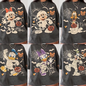 Könnte beinhalten: Ein anthrazitfarbenes T-Shirt mit einem Grafikdruck einer Disney-Figur, die in Mumienbandagen gewickelt ist und einen Kürbis hält. Das Design enthält Spinnennetze, Fledermäuse und Zuckermais, sowie den Namen der Figur in Schreibschrift.