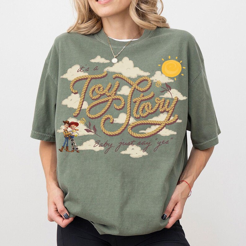 Op de afbeelding: Een olijfgroen t-shirt met een grafische print van de tekst "it's a Toy Story" geschreven in een touwstijl lettertype, vergezeld van illustraties van Woody en Jessie, witte wolken, een gele zon en de tekst "Baby, just say yes".