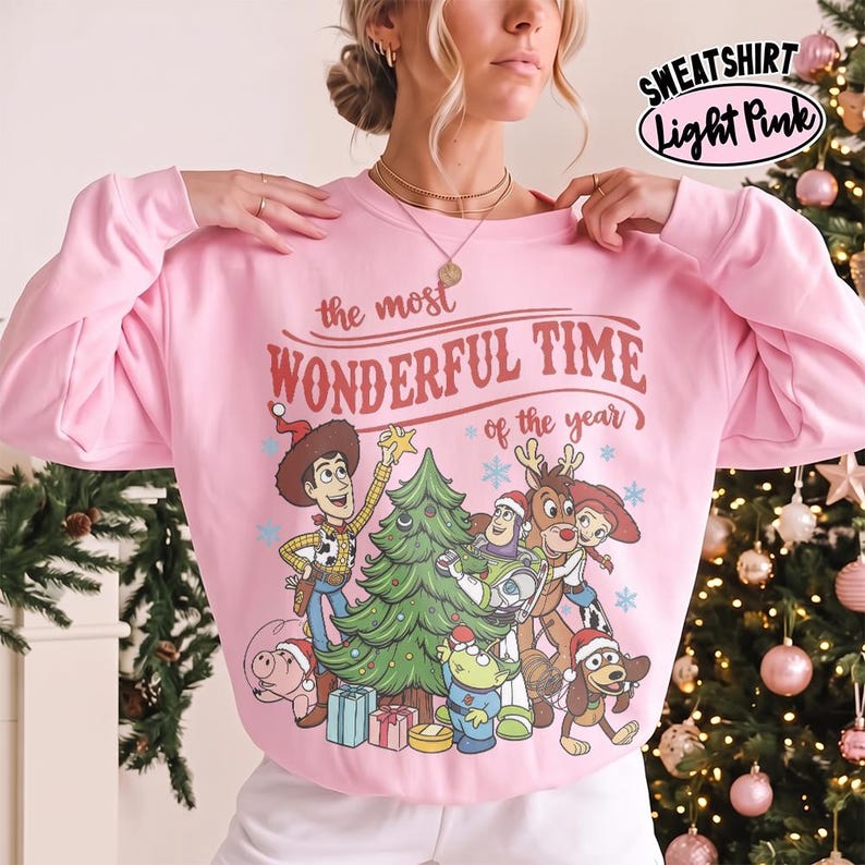May include: A light pink long-sleeve sweatshirt featuring a graphic print of various cartoon characters decorating a Christmas tree. The text reads, "the most WONDERFUL TIME of the year." The design includes Woody, Buzz Lightyear, and other characters wearing Santa hats.