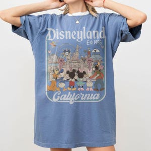 Puede incluir: Camiseta azul con un gr&aacute;fico de Disneyland. El gr&aacute;fico presenta a Mickey y Minnie Mouse, el Pato Donald, Goofy y otros personajes frente a un castillo y una noria. El texto "Disneyland Est. 1955 California" est&aacute; impreso en la parte delantera.
