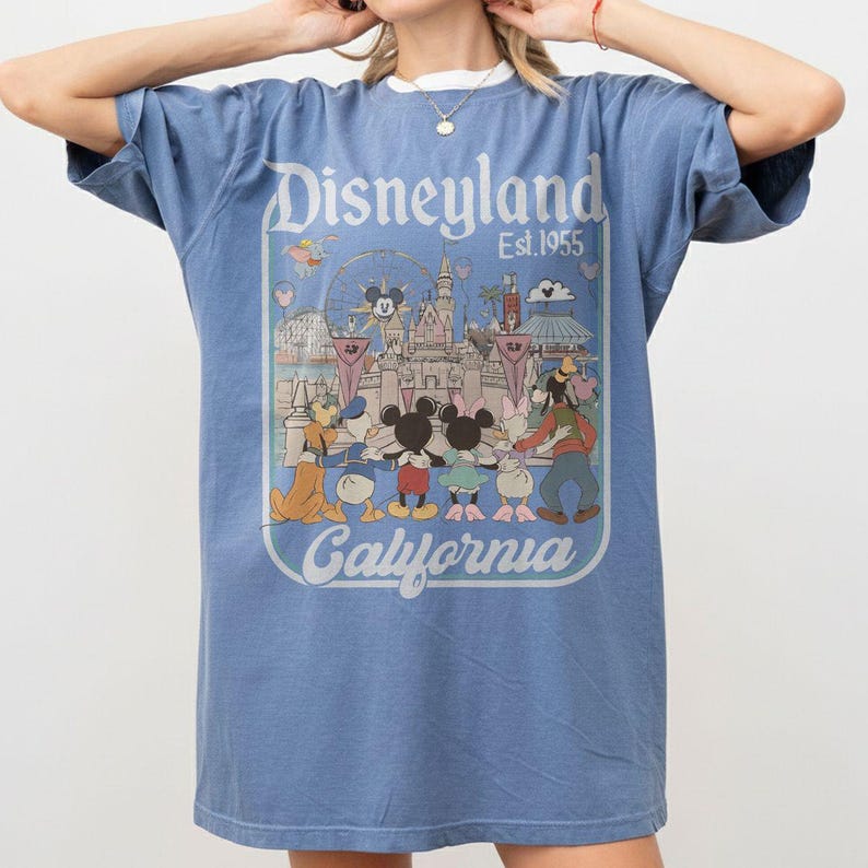 Puede incluir: Camiseta azul con un gr&aacute;fico de Disneyland. El gr&aacute;fico presenta a Mickey y Minnie Mouse, el Pato Donald, Goofy y otros personajes frente a un castillo y una noria. El texto "Disneyland Est. 1955 California" est&aacute; impreso en la parte delantera.