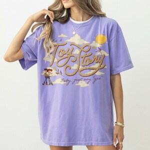 Op de afbeelding: Een lichtpaars t-shirt met korte mouwen en een grafische print van wolken, een gele zon en de tekst "It's a Toy Story" in een touwachtig lettertype. Onder de tekst toont het t-shirt kleine illustraties van de personages Woody en Jessie.