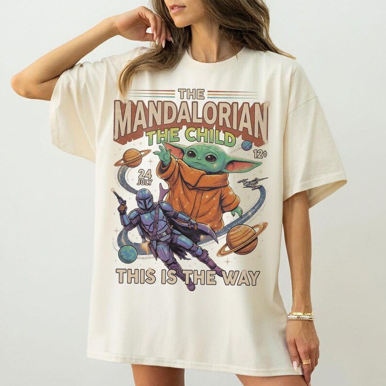 Op de afbeelding: Een off-white t-shirt met korte mouwen en een grafische print met de tekst The Mandalorian The Child en This Is The Way. Het ontwerp bevat een groen buitenaards personage, een figuur in blauw harnas, planeten en sterren in stripstijl.