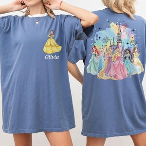 May include: Blue t-shirt featuring a princess graphic on the front and back. The front displays a yellow gown and the name "Olivia". The back showcases a castle and several princesses in colorful gowns.