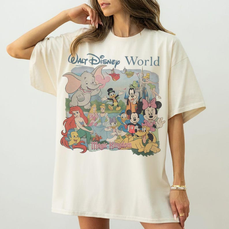 Puede incluir: Una camiseta de manga corta color blanco hueso con un gran estampado gr&aacute;fico de Walt Disney World Magic Kingdom que incluye varios personajes animados como Dumbo, Ariel, Cenicienta, Mickey Mouse, Minnie Mouse, Donald y Goofy.