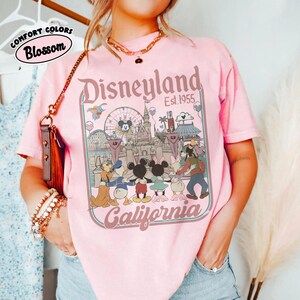 Puede incluir: Camiseta rosa claro con un gr&aacute;fico de Disneyland. El gr&aacute;fico presenta el logotipo de Disneyland, el texto "Est. 1955" y personajes de dibujos animados. La camiseta tambi&eacute;n tiene el texto "Comfort Colors Blossom" en un c&iacute;rculo.