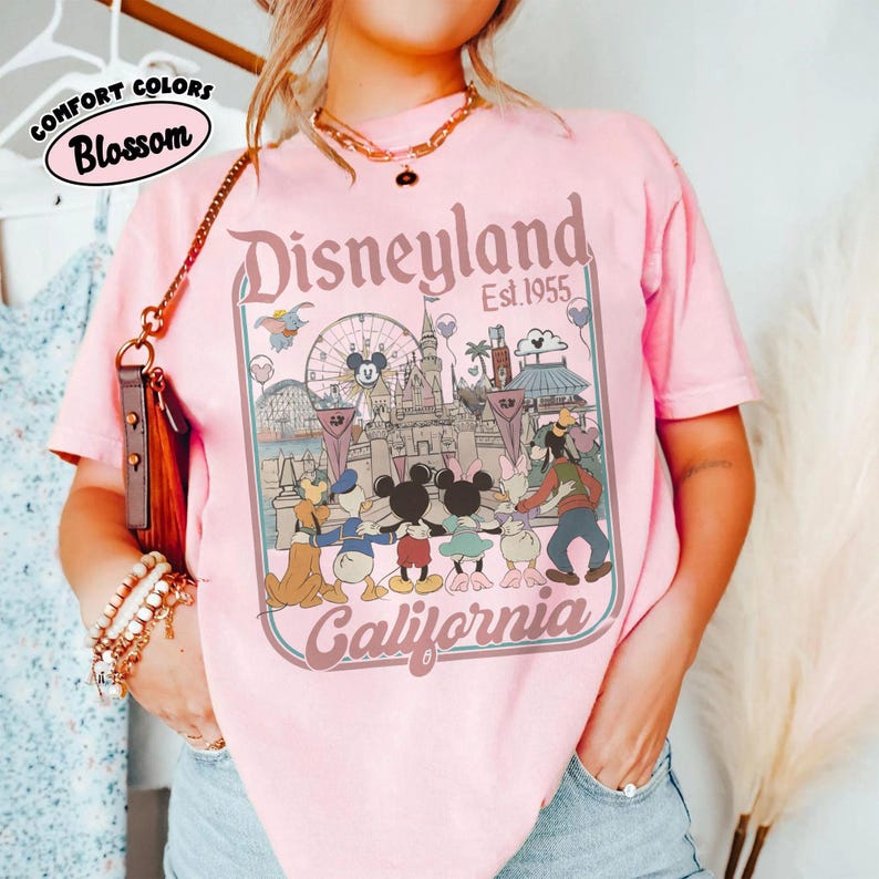 Puede incluir: Camiseta rosa claro con un gr&aacute;fico de Disneyland. El gr&aacute;fico presenta el logotipo de Disneyland, el texto "Est. 1955" y personajes de dibujos animados. La camiseta tambi&eacute;n tiene el texto "Comfort Colors Blossom" en un c&iacute;rculo.