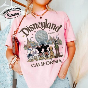 May include: Pink t-shirt with a Disneyland graphic. The design features the Disneyland logo, the date "Est.1955", and cartoon characters like Mickey and Minnie Mouse. The word "CALIFORNIA" is printed below the characters.