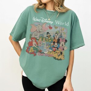 Puede incluir: Una camiseta de manga corta color verde salvia con un estampado gr&aacute;fico de varios personajes de Disney, incluidos Dumbo, Ariel, Cenicienta, el Pato Donald, Goofy, Mickey Mouse, Minnie Mouse y Pluto, con el texto Walt Disney World Magic Kingdom.