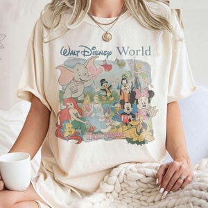 Puede incluir: Una camiseta de manga corta color crema con un estampado gr&aacute;fico de estilo vintage de Walt Disney World y Magic Kingdom con varios personajes, incluidos Dumbo, Ariel, Cenicienta, Mickey Mouse, Minnie Mouse, Pluto y Campanilla.