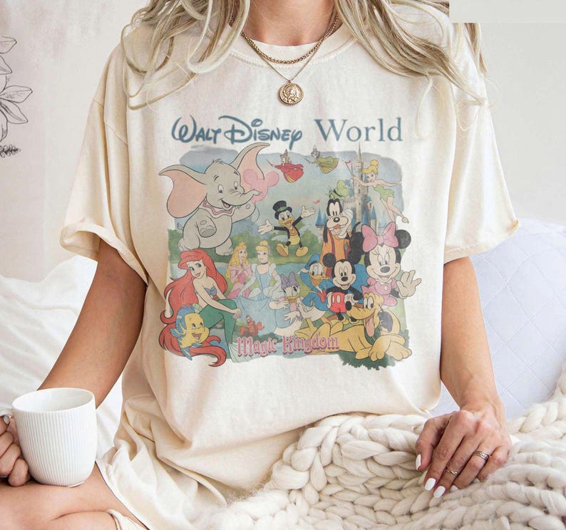 Puede incluir: Una camiseta de manga corta color crema con un estampado gr&aacute;fico de estilo vintage de Walt Disney World y Magic Kingdom con varios personajes, incluidos Dumbo, Ariel, Cenicienta, Mickey Mouse, Minnie Mouse, Pluto y Campanilla.