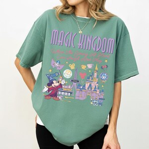 May include: A sage green short-sleeve t-shirt featuring a graphic print with the text "MAGIC KINGDOM" and "Where the Young at Heart Can Laugh and Play". The design includes a wizard Mickey Mouse, a castle, and various small park-themed icons.