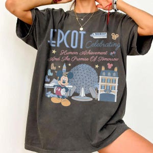 May include: A charcoal gray short-sleeve t-shirt featuring a graphic print that reads EPCOT Celebrating Human Achievement And The Promise Of Tomorrow. The design includes Mickey Mouse, the Spaceship Earth structure, and a monorail train.