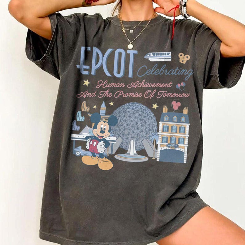 May include: A charcoal gray short-sleeve t-shirt featuring a graphic print that reads EPCOT Celebrating Human Achievement And The Promise Of Tomorrow. The design includes Mickey Mouse, the Spaceship Earth structure, and a monorail train.