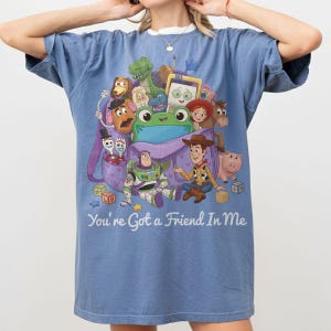 May include: A blue short-sleeve t-shirt featuring a graphic print of various animated characters from Toy Story emerging from a purple backpack, with the text "You're Got a Friend In Me" printed in white script lettering at the bottom of the design.